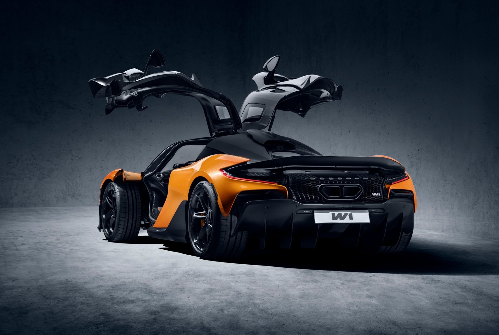 Rear three-quarter view of an orange and black McLaren W1 with its doors open upwards, in a dark studio